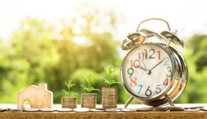 A clock sits next to three stacks of coins and a small house, representative of property management SEO as explained in this blog