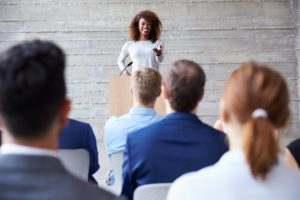 A woman speaks at a podium in front of an audience, representing the NARPM Women's Leadership Council's next event, which is aimed at providing public speaking skills to women in property management