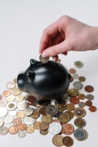 A hand puts coins into a black piggy bank, which sits on top of a pile of coins.