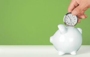 Someone drops a clock into a white piggy bank in front of a green background.