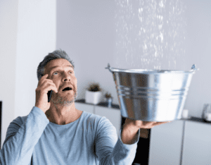 man on phone, holding up bucket to catch water falling from ceiling