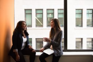Two women laugh and have a conversation next to a window - an example of encouraging referrals by building relationships, an important part of any referral program.