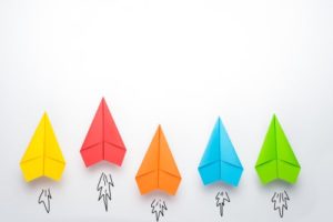 A series of different colored paper airplanes on a white background, with one pulling ahead into the lead, representative of a property management company using Realtor referrals to stay ahead of the competition.