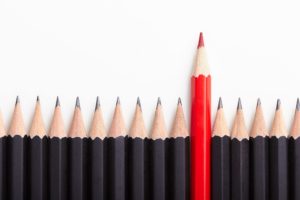 A red colored pencil is pushed above a line of black pencils, standing out from the crowd.
