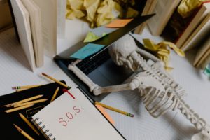 A fake skeleton lays with its head on a laptop covered in sticky notes. Next to it is a notebook with S.O.S. written on it. It's surrounded by books, papers, and scattered pencils.