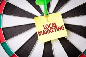 A sticky note with the words "local marketing" is pinned by a dart to a striped dart board.