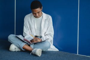 A young adult sits cross-legged in front of a blue wall. They ave a notebook on their lap in which they are writing.