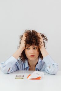 Feminine-presenting person with curly hair sits in front of a table with money and bills, looking worried.